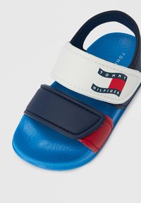 Niebieska klapka z teksturowaną podeszwą, wyposażona w biało-granatowy pasek z czerwonym akcentem i logo "TOMMY HILFIGER".