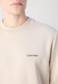 Bežová mikina so zapínaním na krk, vyrobená z hladkej tkaniny, s malým čiernym logom "Calvin Klein" na ľavej hrudi.