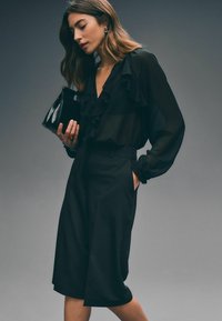Sort gennemsigtig bluse med rynke-detaljer, parret med sorte højtaljede culottes. Modellen holder en lille sort håndtaske og bærer guldøreringe.