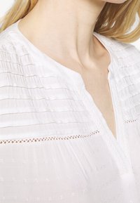 Blouse blanche avec plis verticaux, encolure en V et délicats accents de dentelle. Tissu léger présentant un motif texturé et un léger éclat.