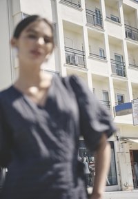 Neaiškus modelio vaizdas tamsiu, tekstūruotu apranga, fone matyti pastatas su balkonais ir viešbučio ženklu.