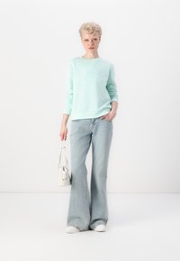 Minta zaļa sporta krekls ar reljefu logo, apvienots ar gaiši ziliem platiem džinsiem. Modelis tur nelielu balto rokassomu, stāvot uz vienkārša fona.