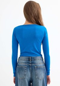 Top de algodón azul de manga larga visto desde la espalda, combinado con unos vaqueros de mezclilla azul claro de cintura alta que tienen dos bolsillos en la parte trasera.