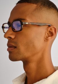 Νεαρός άνδρας με κοντά μαλλιά που φοράει γυαλιά μπλοκαρίσματος μπλε φωτός με σκούρο πλαίσιο τύπου χελώνας και λευκό πουκάμισο με γιακά, κοιτάζοντας προς τα αριστερά με ουδέτερο φόντο.