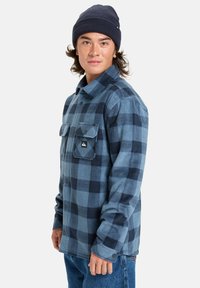 Camisa de franela a cuadros azul y marino con dos bolsillos en el pecho y un parche negro con el logo; combinada con un gorro beanie marino; corte relajado.