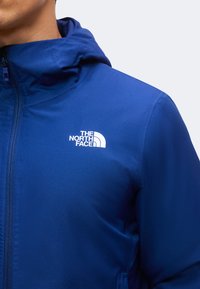 Gros plan d’une personne portant une veste bleue zippée avec une capuche et un logo blanc The North Face sur la poitrine.