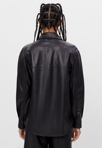 Person med flätat hår bär en långärmad svart läderskjorta, sedd bakifrån mot en enkel ljus bakgrund.