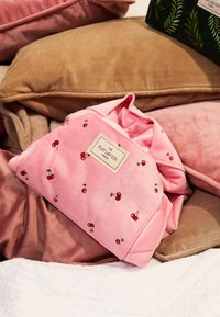 Manta rosa con bordado de cerezas, textura suave y etiqueta que dice "THE FLAT LAY CO. LONDON", colocada entre cojines de tonos neutros.