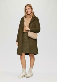 Olijfgroene wolblendjas, beige gebreide coltrui, olijfgroene rok, beige schoudertas en witte veterschoenen met dikke zolen.