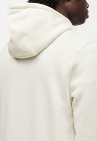 Φούτερ με κουκούλα σε εκρού βαμβακε ρό μείγμα, με απαλή υφή, χαλαρό σχέδιο και ορατές ραφές στην κουκούλα.