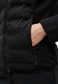 Χέρι εισάγεται στην πλαϊνή τσέπη με φερμουάρ ενός μαύρου επενδυμένου μπουφάν που φοριέται πάνω από ένα μακρυμάνικο μαύρο πουκάμισο.