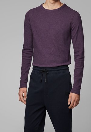 Homme portant un pull prune ajusté à manches longues, rentré dans un pantalon noir à cordon, sur un fond gris uni.