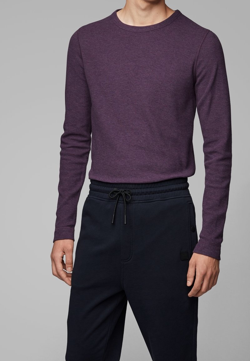 Homme portant un pull prune ajusté à manches longues, rentré dans un pantalon noir à cordon, sur un fond gris uni.