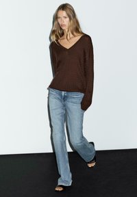Brun strikket sweater med V-hals, kombineret med lyseblå udvidede jeans og sorte hælede sandaler. Ensfarvet baggrund, model går.