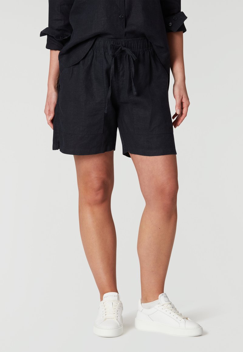 Pantalones cortos de lino azul oscuro con cintura elástica y cordón, que cuentan con dos bolsillos laterales y un ajuste relajado. Combinados con zapatillas blancas.