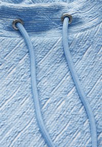 Ljusblå hoodie med texturerat tyg, försedd med två matchande dragsnören som går genom silverfärgade öglor. Texturerad yta med subtila mönster.