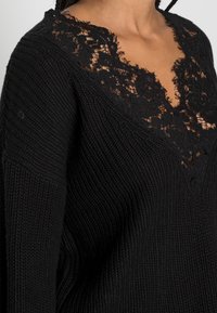 Pull en maille noire avec un décolleté en V profond orné de délicates dentelles noires. La texture est côtelée, mettant en valeur une allure douce et confortable.