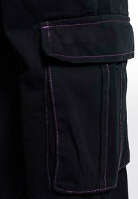 Pantalons cargo noirs avec des coutures violettes contrastantes, plusieurs poches latérales et une texture de tissu durable. Zoom sur le détail de la poche gauche.