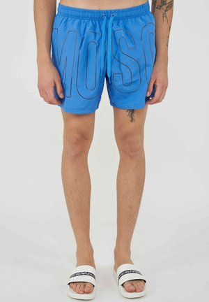 Homme portant un short de bain bleu vif avec cordon, de grandes lettres en contour et des sandales blanches à enfiler avec un logo noir.