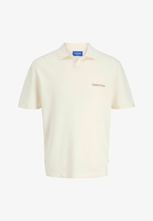 Dospievajúci s kučeravými vlasmi stojí a pozerá sa doprava, oblečený v krémovej polokošeli s krátkym rukávom, svetlošedých nohaviciach a béžovo-bielych teniskách na jednoduchom pozadí.