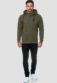 Chupa de sudadera verde oliva con cremallera, fabricada con tejido texturizado, con cordones ajustables y múltiples bolsillos con cremallera, combinada con jeans negros y zapatillas blancas.