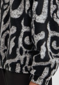 Zwarte en witte gewatteerde fleece jas met een abstract gedraaid patroon, gedragen over zwarte broek, waarbij de onderzijde van de mouw en de zijkant zichtbaar zijn.
