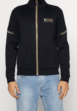 Czarna kurtka na suwak z złotym logo "BOSS HUGO BOSS" na piersi oraz złotymi akcentami na rękawach, noszona z jasnoniebieskimi dżinsami.