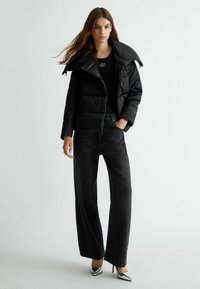 Giacca a vento trapuntata nera con colletto alto, indossata sopra un top nero e abbinata a jeans in denim scuro a gamba larga e tacchi argentati.