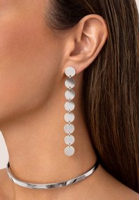 De longues boucles d'oreilles en argent avec des disques circulaires reliés par de petites sections de chaîne, présentant une texture lisse et brillante. Un collier ras du cou en argent complète les boucles d'oreilles.