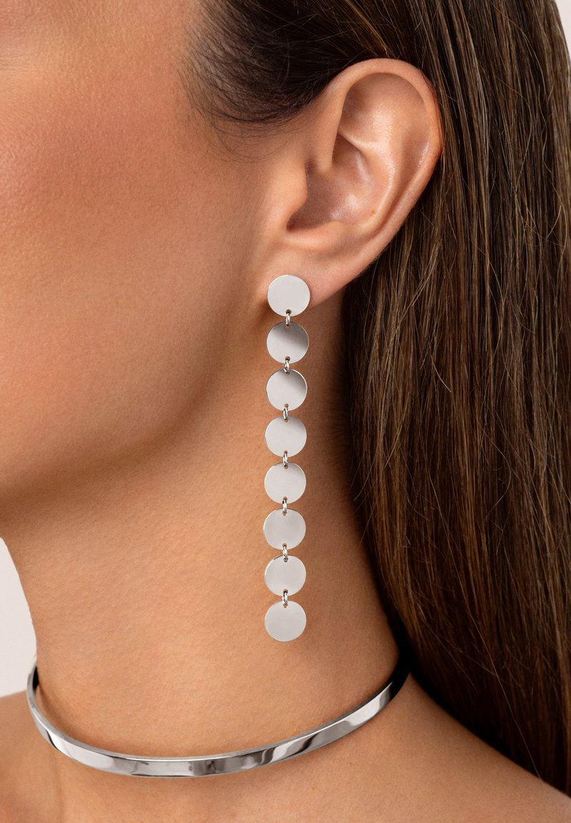 De longues boucles d'oreilles en argent avec des disques circulaires reliés par de petites sections de chaîne, présentant une texture lisse et brillante. Un collier ras du cou en argent complète les boucles d'oreilles.