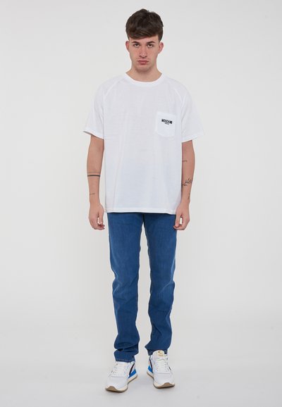 T-shirt bianca oversize con tasca sul petto e logo nero, abbinata a jeans in denim blu e sneakers bianche con dettagli blu.