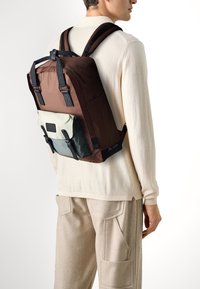 Hnědý látkový batoh s kontrastními světle zelenými a tmavě modrými předními kapsami. Má nastavitelné popruhy a texturovaný povrch.