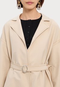 Beige Mantel mit Gürtel, großem Kragen und silbernem, rundem Metallverschluss, kombiniert mit einem schwarzen, gerippten Oberteil mit sechs Knöpfen. Glatte Textur.