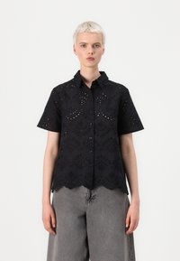 Blusa nera a maniche corte con intricati motivi a occhio di pernice, orlo scallop e chiusura frontale con bottoni. Realizzata in tessuto leggero.