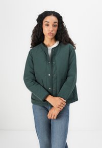 Veste verte matelassée avec un col rond, des fermetures à pression, deux poches latérales et une coupe décontractée, associée à un jean bleu.