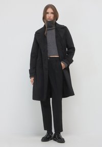 Cappotto lungo di lana nera con colletto, maniche lunghe e bottoni frontali, abbinato a un dolcevita grigio a coste e pantaloni neri sartoriali.