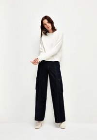 Bílý svetr s květinovou výšivkou, kalhoty z tmavého denimu s vysokým pasem a béžové kotníkové boty na blokovém podpatku. Jednoduchý, moderní design.