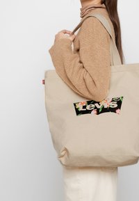 Beige kanvaskasse med en svart blomsterlogotyp i en patch som läser "Levi's." Har robusta handtag och en enkel, ostrukturerad siluett.