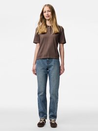 Sieviete ar gariem blondiem matiem, tērpusies brūnā īsroku polo krekliņā, zilos džinsos un brunčos brūnās sporta apavos, stāvot pret balto fonu.