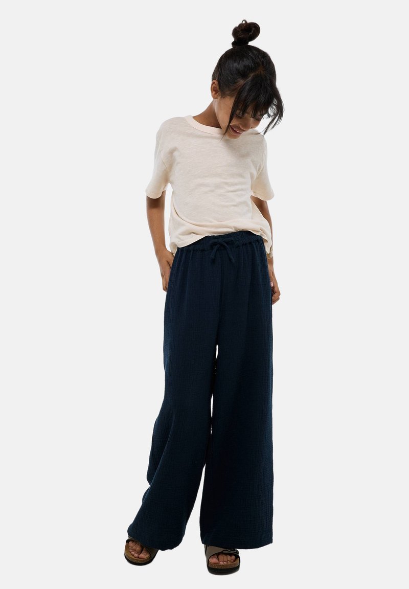 Chemise courte beige clair à manches courtes et pantalons larges bleu marine en tissu texturé. Les pantalons sont dotés d'une ceinture élastique avec cordon de serrage. Sandales décontractées.