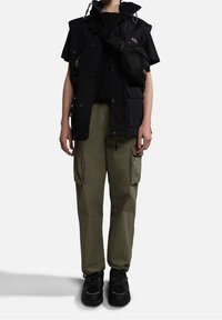 Svart kortärmad väst med flera fickor över en svart topp, i kombination med olivgröna cargos och svarta sneakers, med en liten axelväska.