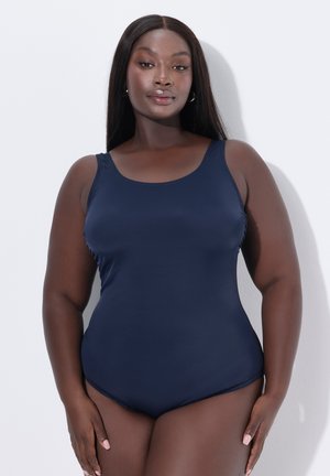 Vrouw met lang steil haar die een mouwloze marineblauwe eendelige badpak draagt tegen een effen lichte achtergrond.