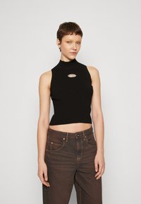 Schwarzes, geripptes Cropped-Top mit hohem Kragen und einem Metallschriftzug vorne, kombiniert mit dunkelbraunen Jeans mit Kontrastnähten.