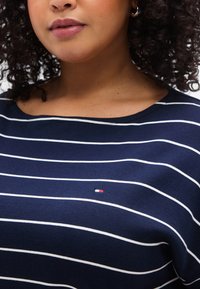 Μπλε σκούρο πουκάμισο με λευκές οριζόντιες ρίγες, στρογγυλή λαιμόκοψη και μικρό λογότυπο σε ταμπέλα με κόκκινες, λευκές και μπλε αποχρώσεις στο στήθος.