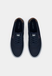 Mornarske platnene superge z belimi gumijastimi podplati, okrogli prsti in šestimi odprtinami za vezalke. Rjavi detajli na peti in blagovna znamka na ježku.