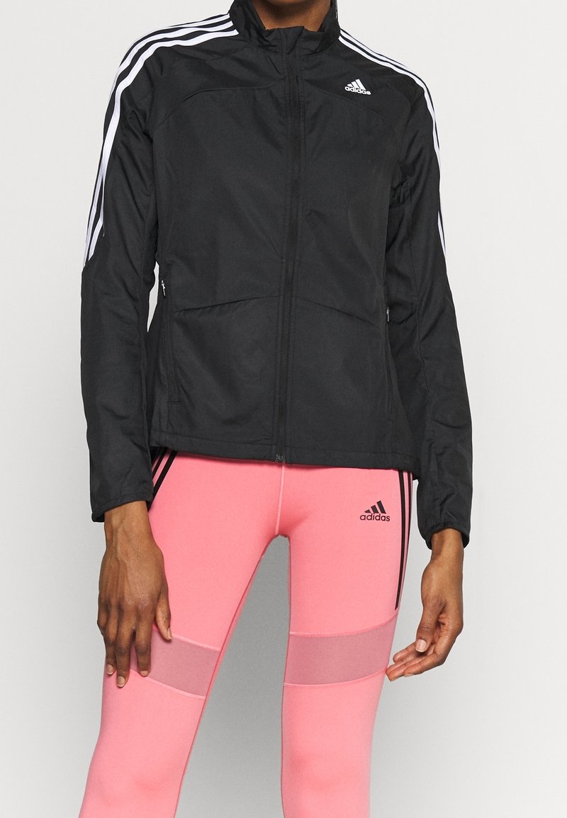 Personne portant une veste Adidas noire avec des bandes blanches et des leggings Adidas roses avec des panneaux en maille sur les cuisses.