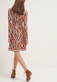 Robe à manches longues avec un motif ondulé aux tons rose, orange et noir, taille cintrée et ourlet à hauteur des genoux. Bottines en daim marron.