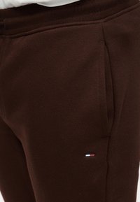 Καφέ φόρμα από απαλό ύφασμα, με ριμπ ζώνη μέσης και πλαϊνές τσέπες, διακοσμημένη με ένα μικρό κεντητό λογότυπο.