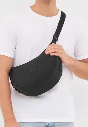 Hombre con camiseta blanca sostiene un bolso cruzado negro en forma de media luna con correa ajustable sobre el pecho.