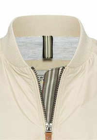 Gros plan sur un col de veste beige avec bordure en tricot texturé, fermeture éclair à moitié fermée avec tirette en tissu rayé, et ruban bouclé à l'intérieur du col.